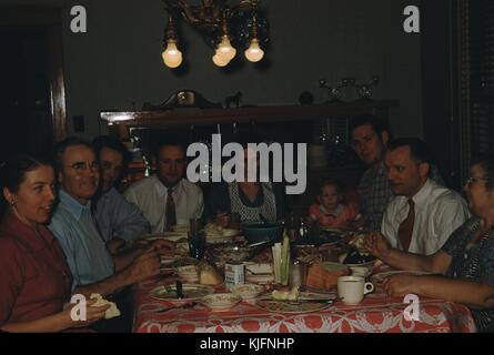 A photograph of a family sitting for dinner together at a table, three generations of the family appear ranging from a toddler to grandparents, the table is covered in plates, food, utensils and condiments, 1952. Stock Photo