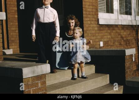 A photograph of a woman and two children posing for a photograph on a concrete and brick porch, all three are wearing formal clothing, the toddler girl is wearing a blue dress and shiny black shoes, the boy wears a bowtie with his button down white shirt that has been tucked in to blue slacks, the mother wears a black top with a blue skirt and a necklace, 1952. Stock Photo