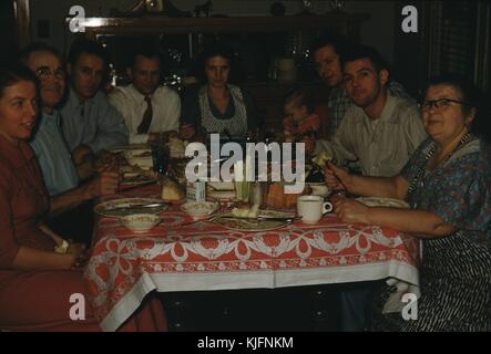 A photograph of a family sitting for dinner together at a table, three generations of the family appear ranging from a toddler to grandparents, the table is covered in plates, food, utensils and condiments, 1952. Stock Photo