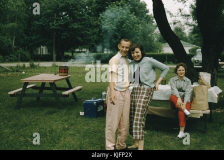 Couple posing for a picture at a park, man is smoking a cigarette, behind them a woman sitting at a picnic table covered with a table cloth, a cooler, and what appear to be bags of groceries, the people all seem to be in 1950s attire, 1952. Stock Photo