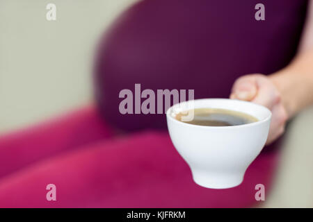 Young pregnant woman sitting in a kitchen and holding  a cup od coffee in a hand. Pros and cons coffee concept. Stock Photo