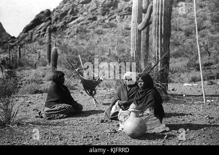 A photograph of three Native American women resting, they are all wearing long dresses with light blankets around their shoulders, they have a jug of water and a basket for collecting food with them, they are resting among the desert foliage during a break from harvesting, 1907. From the New York Public Library. Stock Photo