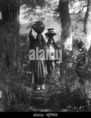 A photograph of two Native American women carrying jugs of water on their heads, they are walking along a path through the forest returning from the river, they wear long flowing dresses and head coverings, Taos, New Mexico, 1905. From the New York Public Library. Stock Photo