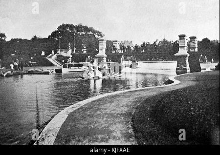 A photograph of the Public Garden, people can be seen traveling on the water in boats while other people watch them from a walking bridge, the water is lined on both sides by sidewalks and grass, the park is located in the heart of the city, it was established in 1837 by Horace Grey, his aim was to make it the first public botanical garden in the country, in 1987 it was declared a National Historic Landmark, Boston, Massachusetts, 1905. Stock Photo