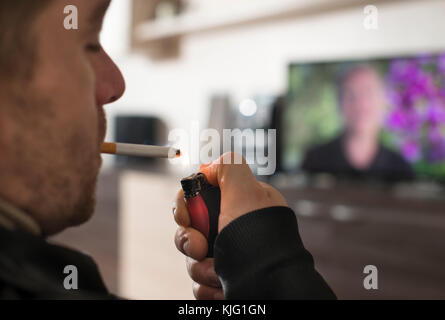 Young man lights a cigarette in front of TV Stock Photo