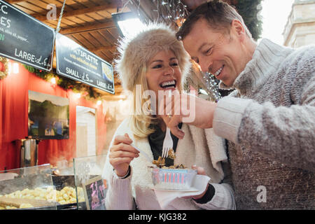 Mature couple are sharing a carton of food they have bought from a french food stall in the christmas market. Stock Photo