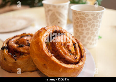 Two rolls with raisins and cinnamon are lying on a white plate on the table against paper glasses of tea. Stock Photo