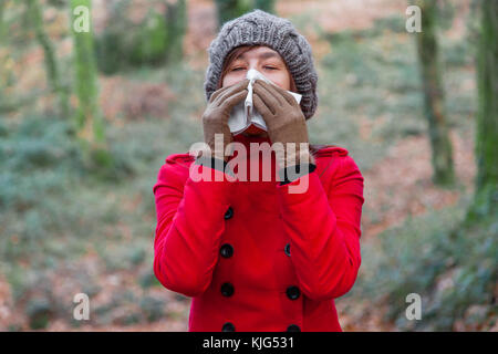 Young woman suffering from cold or flu blowing nose or sneezing on white paper handkerchief in forest wearing a red long coat or overcoat and beanie
