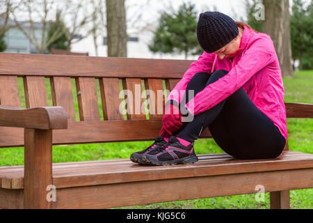 Female athlete tired or depressed resting on a bench on a cold winter day on the training track of an urban park. Young woman wearing pink windbreaker Stock Photo