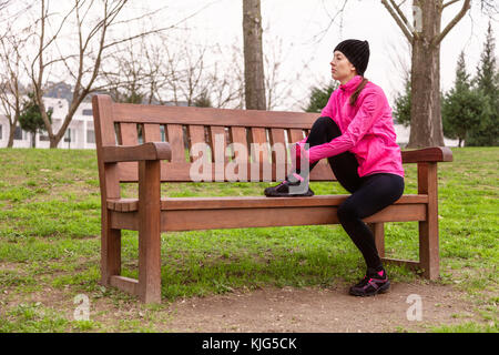 Female athlete tired or depressed resting on a bench on a cold winter day on the training track of an urban park. Young woman wearing pink windbreaker Stock Photo