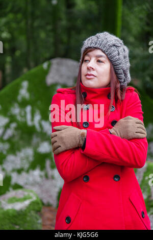 Young woman lost on a forest shivering with cold and embracing or holding herself, wearing a red long coat or overcoat, a beanie and gloves during fal Stock Photo