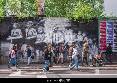 Berlin wall, people walk past a mural depicting Hitler Youth in stylised form painted on the Berlin Wall, East Side Gallery, Friedrichshain, Berlin. Stock Photo