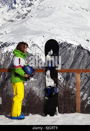 Young snowboarder with helmet in hands and snowboard on viewpoint in winter mountain at sunny day. Caucasus Mountains. Hatsvali, Svaneti region of Geo Stock Photo