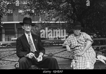 A man and a woman sitting on a metal park bench, the man is wearing a suit and tie along with a hat, the woman is wearing a patterned dress and a hat, a portion of Jackson Square can be seen behind them with cars and a building just outside of the park, New Orleans, Louisiana, 1935. From the New York Public Library. Stock Photo