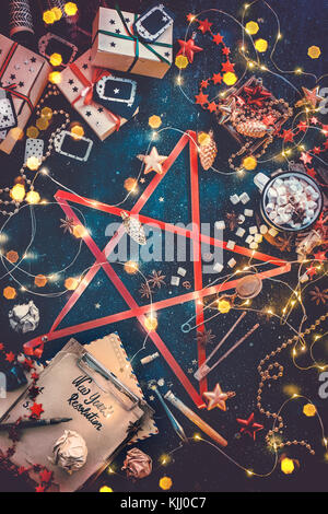 Star of Christmas essentials. Decorations, gift boxes, fairy lights, new year resolution notebook and hot chocolate connected with a red ribbon into a Stock Photo