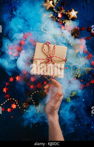 Christmas surprise concept, a gift box with dence steam and a reaching hand on a dark background with decorations and stars. New year present vivid st