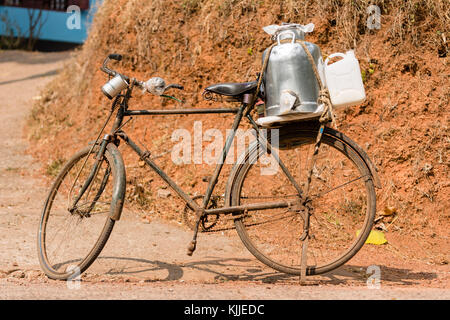 An old and rusty bicycle stands on the road side in Kerala, India. It is used by a milkman for deliveries and features a tin gallon and serving cups. Stock Photo