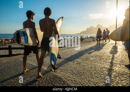 RIO DE JANEIRO - MARCH 24, 2017: Sunset silhouettes of two young surfers walking with surfboards at Arpoador with two brothers mountains in the backgr