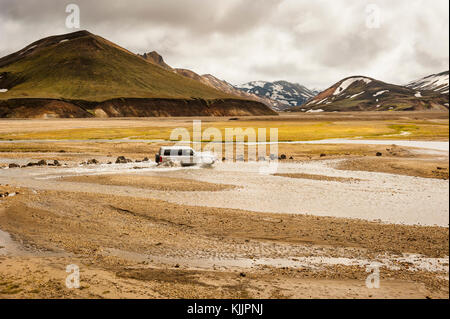 4x4 vehicle, SUV crossing a stream with Landmannalaugar colourful mountains still covered with snow, beginning of Summer, Iceland Highlands, Europe. Stock Photo