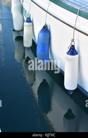 White and blue plastic fenders hanging on ropes along white ship hull. Stock Photo