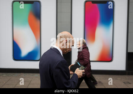 A man uses his handset while walking past an ad for the new Apple iPhone X outside a branch of Carphone Warehouse, on 22nd November 2017, in London England. Stock Photo