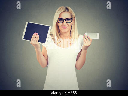 Perplexed woman in glasses shrugging shoulders uncertain what to choose tablet or smartphone Stock Photo