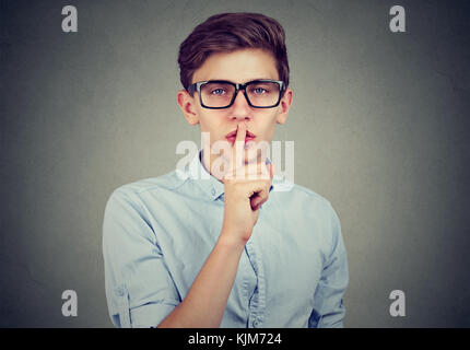 Secret guy. Man saying hush be quiet with finger on lips gesture looking at camera isolated on gray wall background. Stock Photo