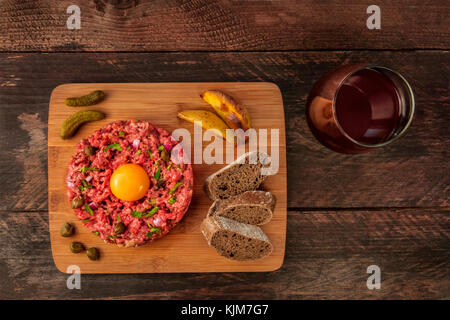 A photo of a steak tartare with a raw egg yolk, gherkins, capers, rye bread, roasted potatoes, a glass of red wine, shot from above on a dark rustic t Stock Photo