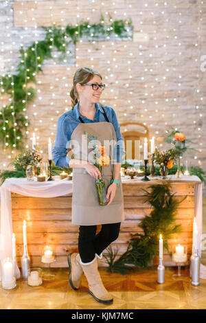 floral design, profession, decoration concept. cute smiling girl dressed in clean apron and comfortable shirt holding vase with orange flowers collected in small but lovely bunch on festive background Stock Photo