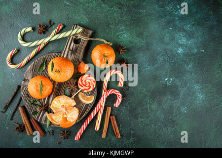 Fresh mandarins with leaves, candy canes and spices - cinnamon, anise and cloves on a dark cutting board on a dark green background. Top view Stock Photo