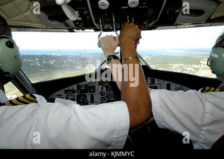 A female co-pilot with hand on throttles and a male captain with arms crossed over in the cockpit of a Winair DHC-6 Twin Otter on final-approach SXM-A Stock Photo