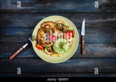 Roasted chicken with vegetables and green mashed potatoes. Fork, knife on a colored wooden background. Top view Stock Photo