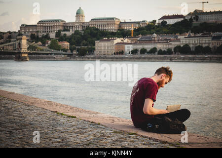 Young man sitting on the Danube river bank reading a book in Budapest (Hungary). June 2017. Landscape format. Stock Photo