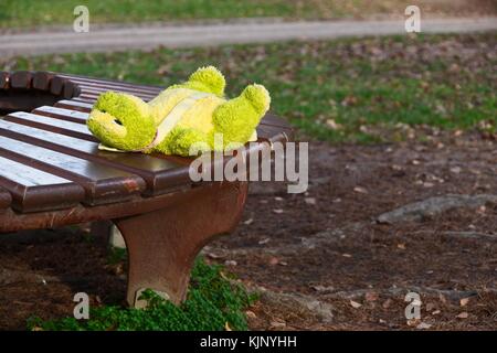 Lonely forgotten soft green frog toy, child backpack, lying on the bench in park waiting for owner, child to return Stock Photo