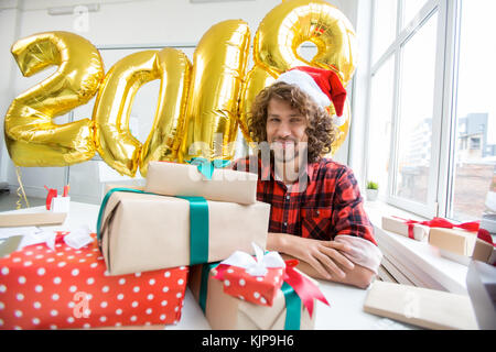 Happy young Santa with stack of gift-boxes in front and golden balloons in form of 2018 behind Stock Photo