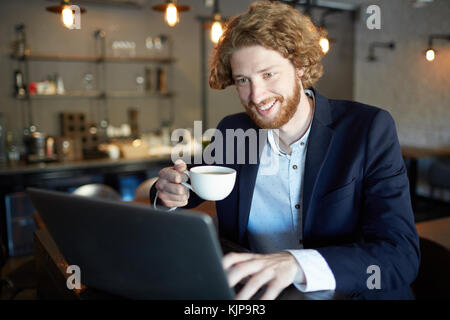 Happy young man with cup of tea working in front of laptop in cafe or watching curious video Stock Photo