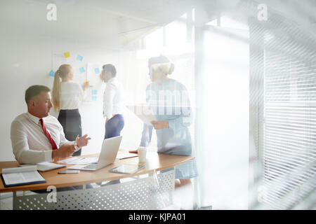 Economists discussing online financial statistics in office on background of two colleagues planning work by whiteboard Stock Photo