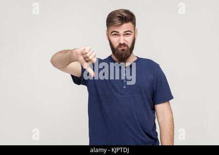Portrait of unsatisfied bearded man with thumbs down and blue t-shirt against gray background. Studio shot. Stock Photo