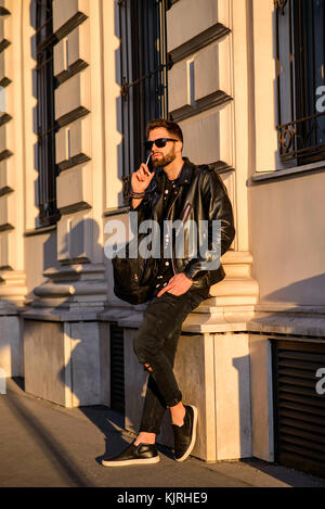 A handsome young hipster man leaning to the wall of a house on a sunny street while talking on his smartphone Stock Photo