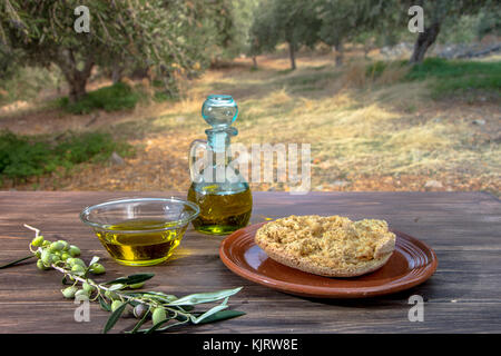 Bowl and bottle with extra virgin olive oil, olives, a fresh branch of olive tree and cretan rusk dakos on wooden table, in an olive tree field at Cre Stock Photo