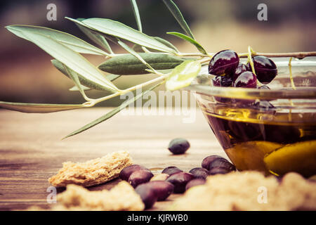 Bowl with extra virgin olive oil, olives, a fresh branch of olive tree and cretan rusk dakos close up on wooden table, Crete, Greece. Stock Photo
