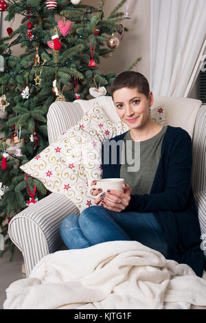 Young beautiful woman with short hair enjoying cup of tea, sitting in front of christmas tree. Authentic family xmas time