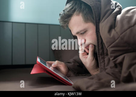 Man is working at night, lying on the sofa in the living room with tablet, covering his head with blanket. Stock Photo