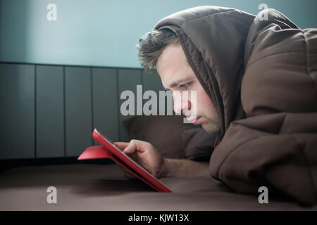 Man is working at night, lying on the sofa in the living room with tablet, covering his head with blanket. Stock Photo