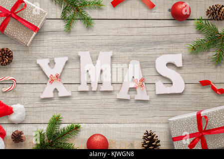 Xmas white letters on wooden table surrounded with Christmas decorations, gifts, fir branches, balls, Santa hat and pinecones. Top view. Stock Photo