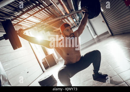 Young muscular man doing overhead squat exercise with barbell on cross training at the garage gym. Stock Photo