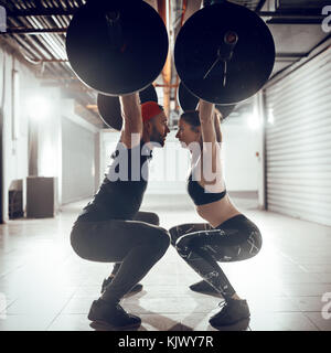 Young muscular couple doing overhead squat exercise with barbell on cross training at the garage gym. Stock Photo