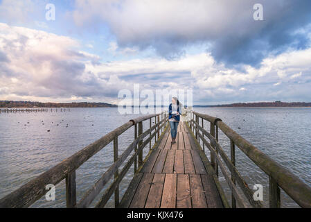 Beautiful young woman smiling and walking alone on a long wooden pontoon, over the lake Chiemsee, enjoying the fresh air and the sunset, in Germany. Stock Photo