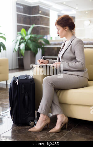 Full length portrait of elegant young businesswoman in hotel lobby waiting and making notes sitting on sofa with suitcases Stock Photo