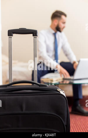 Blurred portrait of handsome bearded businessman using laptop working in hotel room, focus on suitcase in foreground Stock Photo